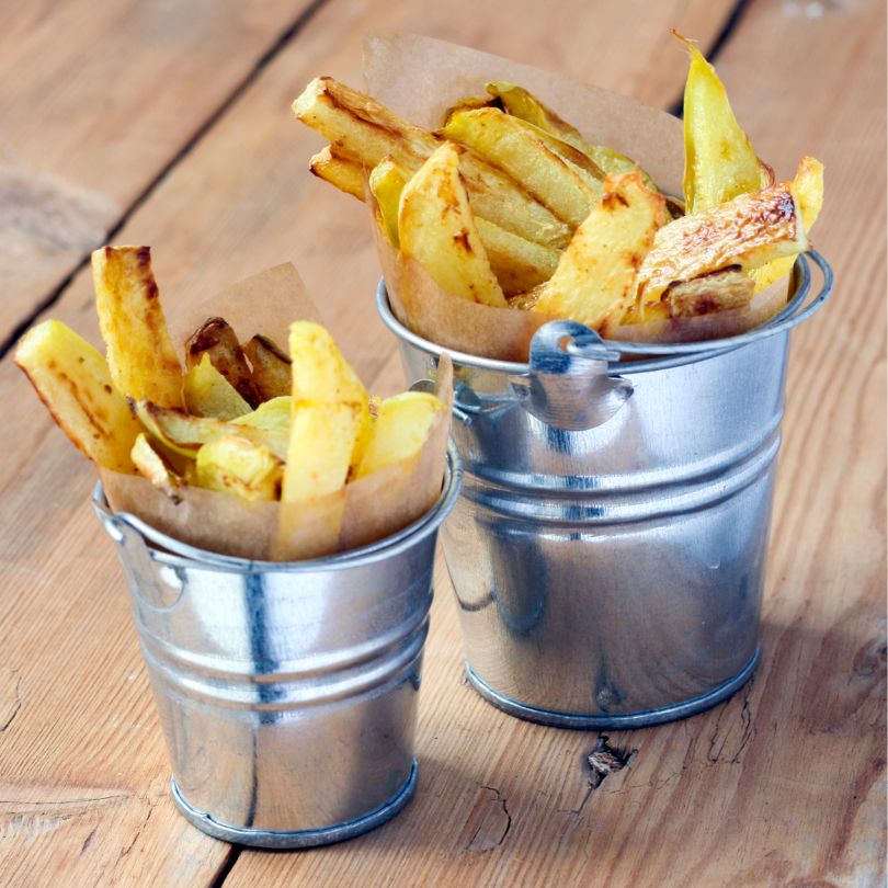 Knusprige ketogene Pommes aus Kohlrabi in zwei kleinen Metalleimern mit Backpapier serviert, goldbraun gebacken und appetitlich angerichtet auf einem rustikalen Holztisch.